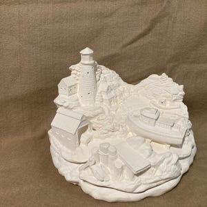 May include: A white ceramic coastal scene sculpture featuring a lighthouse, buildings, and a boat. The detailed design includes textured surfaces and a rounded base, creating a miniature seaside village.