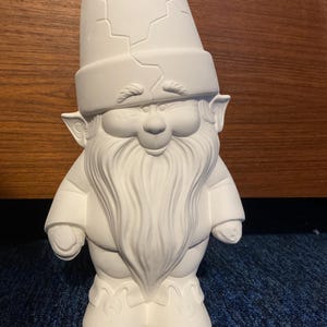 May include: White ceramic gnome figurine with a cracked hat, a long beard, and a smiling face. The gnome is standing with its arms at its sides.