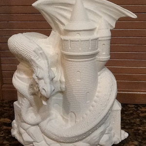 May include: A white ceramic dragon figurine with a castle on its back. The dragon is curled around the castle, and its wings are spread out. The figurine is on a wooden base.