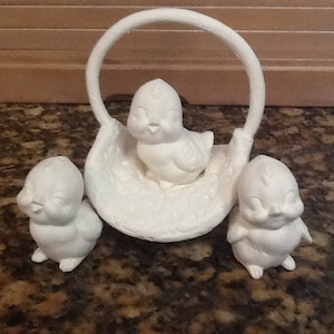 May include: Three white ceramic birds, two standing on the ground and one sitting inside a white ceramic basket. The basket has a round handle.