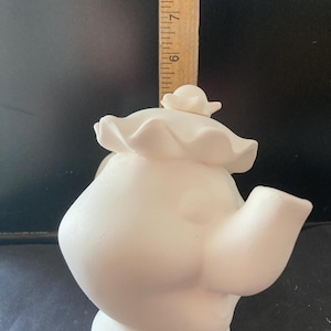 Puede incluir: Una tetera de cerámica blanca con un diseño caprichoso. La tetera tiene un cuerpo redondo, un pico y un asa. Se coloca una regla de madera junto a la tetera. La tetera no está pintada y está lista para personalizar.