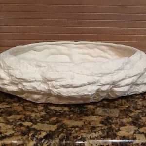 Puede incluir: Maceta de cerámica blanca con forma de roca con una parte superior redondeada en forma de cuenco. La maceta está diseñada para parecer una formación rocosa natural.