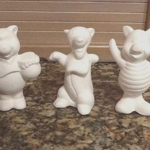 以下が含まれることがあります： 漫画のキャラクターの未塗装のセラミックフィギュア3体。最初のフィギュアは蜂蜜の壺を持っているクマです。2番目のフィギュアは腕を広げたクマです。3番目のフィギュアは手を振っている豚です。すべてのフィギュアは白です。