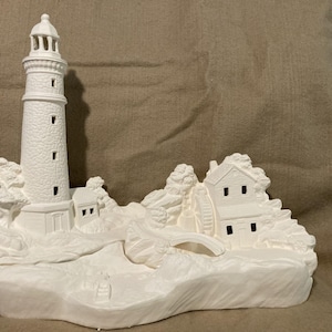 May include: A white ceramic lighthouse sculpture with a detailed design. The sculpture features a tall lighthouse, a small house, and a bridge, all set on a rocky base. The piece is unpainted and ready for customization.