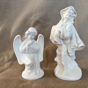 Può includere: Due statuette in ceramica bianca. Una è un angelo in ginocchio con le mani giunte e grandi ali. L'altra è una figura in piedi in abiti fluenti, che tiene un fascio. Entrambe sono non verniciate e pronte per la personalizzazione.