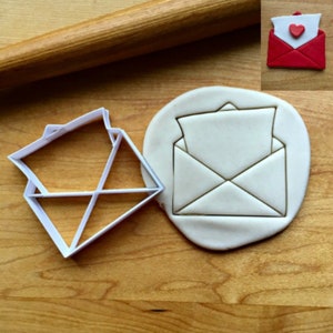 May include: A white plastic cookie cutter in the shape of an envelope with a folded letter inside. A piece of white fondant is pressed into the shape of the envelope using the cookie cutter.