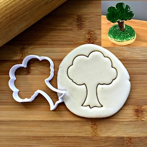 May include: A white plastic cookie cutter shaped like a tree with a rounded crown and a trunk. The cutter is on a wooden surface next to a piece of white dough with the tree shape cut out.
