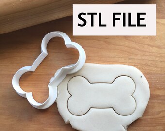 May include: A white plastic dog bone-shaped cookie cutter next to a piece of dough with the bone shape cut out. The text "STL FILE" is in a white rectangle in the upper right corner. The background is a wooden surface.