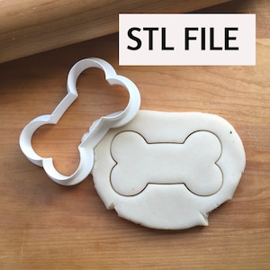 May include: A white plastic dog bone-shaped cookie cutter next to a piece of dough with the bone shape cut out. The text "STL FILE" is in a white rectangle in the upper right corner. The background is a wooden surface.