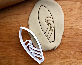 Könnte beinhalten: Weißer Plastik-Ausstecher in Form eines Surfboards mit Wellendesign. Der Ausstecher liegt auf einem Stück Keksteig.
