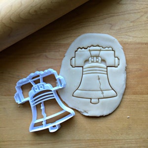 May include: A white plastic cookie cutter shaped like the Liberty Bell. The cutter is next to a piece of unbaked cookie dough with the Liberty Bell shape pressed into it.