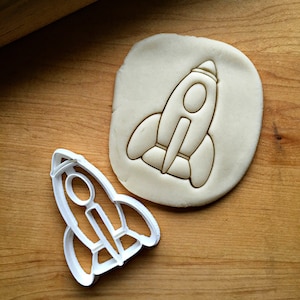 May include: A white plastic cookie cutter shaped like a rocket ship. The cookie cutter is on a wooden surface with a piece of dough that has been cut out with the cookie cutter.