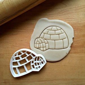 May include: A white plastic cookie cutter in the shape of an igloo. The cutter is shown on a wooden surface with a piece of dough that has been cut out with the cutter.