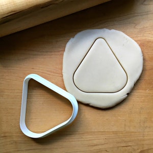 May include: A light blue plastic cookie cutter in the shape of a triangle next to a piece of dough with a triangle cut out. A wooden rolling pin is in the background. The dough and cutter are on a wooden surface.