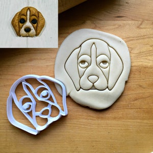 May include: A white plastic cookie cutter in the shape of a beagle dog's face, next to a cut-out cookie and a finished cookie. The cookie cutter is on a wooden surface.