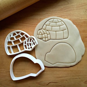 May include: White plastic igloo-shaped cookie cutters next to a cut-out igloo shape in dough. The cutters are in two pieces, one for the main structure and one for the entrance. The dough shows the igloo outline and details.