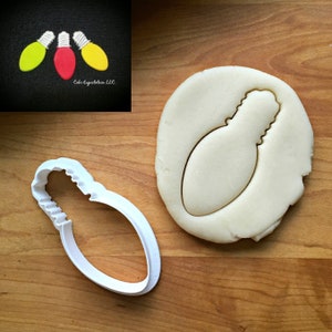 May include: A white plastic cookie cutter shaped like a light bulb. The cutter is shown on a wooden surface with a piece of dough that has been cut out using the cutter.