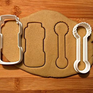 May include: A close-up shot of a piece of dough with three cookie cutters. The cookie cutters are white plastic and are in the shape of a bottle, a spoon, and a bottle with a scalloped top. The dough is light brown and sits on a wooden surface.