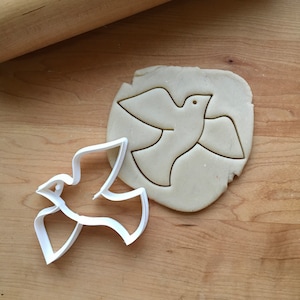 May include: White plastic cookie cutter in the shape of a dove, with a dove outline cut into a piece of raw cookie dough.
