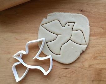 Può includere: Tagliapasta in plastica bianca a forma di colomba, con un contorno di colomba tagliato in un impasto per biscotti crudo.