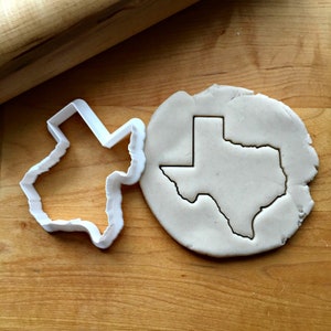 May include: A white plastic cookie cutter in the shape of the state of Texas, with a piece of cookie dough cut out using the cutter.