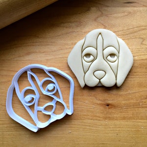 May include: A dog-shaped cookie cutter and a baked cookie. The cookie cutter is white plastic, and the cookie is beige with detailed facial features. The rolling pin is visible in the background. The cookie and cutter are on a wooden surface.