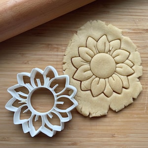 May include: A white plastic cookie cutter shaped like a sunflower, next to a cookie that has been cut out using the cutter. The cookie is a light brown color.