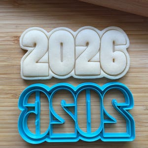 May include: A cookie cutter and a baked cookie, both shaped with the numbers 2026. The cookie cutter is bright blue, and the cookie is a light beige color. The items are on a wooden surface.