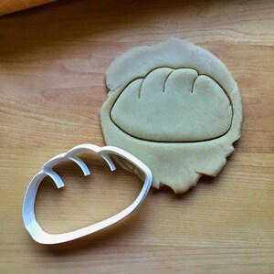 May include: A white plastic cookie cutter shaped like a loaf of bread. The cutter is on a wooden surface next to a piece of dough that has been cut out with the cutter.