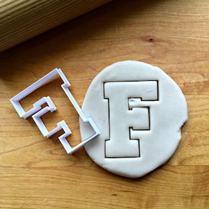 May include: White plastic cookie cutter in the shape of the letter F. The cutter is next to a piece of white modeling clay with the letter F cut out.