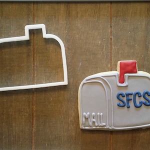 May include: A mailbox-shaped cookie and a white plastic cookie cutter sit on a wooden surface. The cookie is gray with blue lettering that reads "SFCS" and "MAIL". A red flag is raised. The cookie cutter is a white outline of a mailbox.