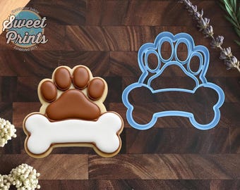 May include: A light blue plastic cookie cutter shaped like a paw print above a bone, alongside a finished cookie decorated with brown icing on the paw print and white icing on the bone, featuring the text Sweet Prints Established 2015.