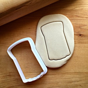 Peut inclure: Un emporte-pièce en plastique blanc en forme de tranche de pain est posé à côté d'un morceau de pâte avec la forme de pain découpée. Un rouleau à pâtisserie en bois est en arrière-plan. Les éléments sont sur une surface en bois.