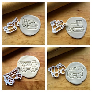 May include: Four cookie cutters in the shape of construction vehicles. The cutters are on a wooden surface with a rolling pin. The cutters are white and the vehicles are a dump truck, a cement mixer, a bulldozer, and an excavator.