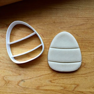 May include: A white plastic egg-shaped cookie cutter with three horizontal bars next to a light-coloured egg-shaped biscuit with three horizontal indentations. The items are on a wooden surface.