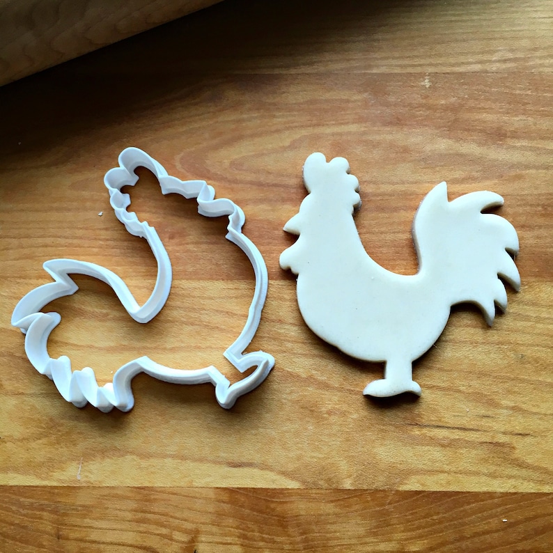 May include: A white plastic rooster-shaped cookie cutter positioned next to a matching rooster-shaped piece of cut dough. The cookie cutter features a detailed outline of a rooster with a prominent comb, beak, and tail feathers, mirroring the dough.