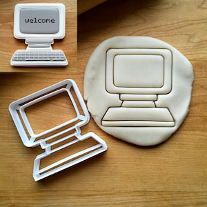 May include: A white computer-shaped cookie cutter and a corresponding computer-shaped cookie with gray icing and the text "welcome" on the screen. The cookie cutter features a classic desktop monitor and keyboard outline design in white plastic.
