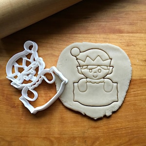 May include: White plastic cookie cutter shaped like an elf holding a sign. The elf has a red hat with a white pom-pom, a white collar, and a smiling face. The sign is blank.