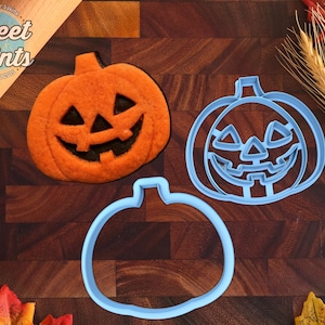 May include: An orange pumpkin-shaped cookie with a jack-o'-lantern face, a light blue pumpkin-shaped cookie cutter with an internal jack-o'-lantern face design, and a light blue pumpkin-shaped cookie cutter outline. The text Sweet Prints is visible.