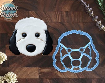 May include: A dog-shaped cookie decorated with white and black icing to resemble a doodle or poodle face, positioned next to a light blue plastic cookie cutter that matches the outline and internal facial features of the dog design.