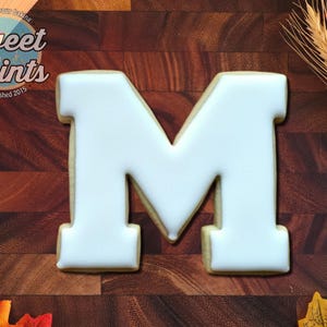 May include: A letter M shaped sugar cookie with smooth white royal icing. The cookie features a block letter design with clean edges. The text Sweet Prints Established 2015 is visible in the upper left corner of the image, along with wheat stalks.