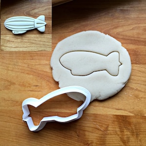 May include: White plastic cookie cutter shaped like an airship or blimp. The cutter is shown on a wooden surface with a piece of dough cut out with the cutter.