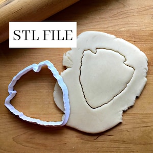 May include: A white plastic cookie cutter in the shape of a shield, with a slightly textured edge, is shown on a wooden surface. The cookie cutter is being used to cut out a shield shape from a piece of dough. The text "STL FILE" is visible in the upper left corner of the image.