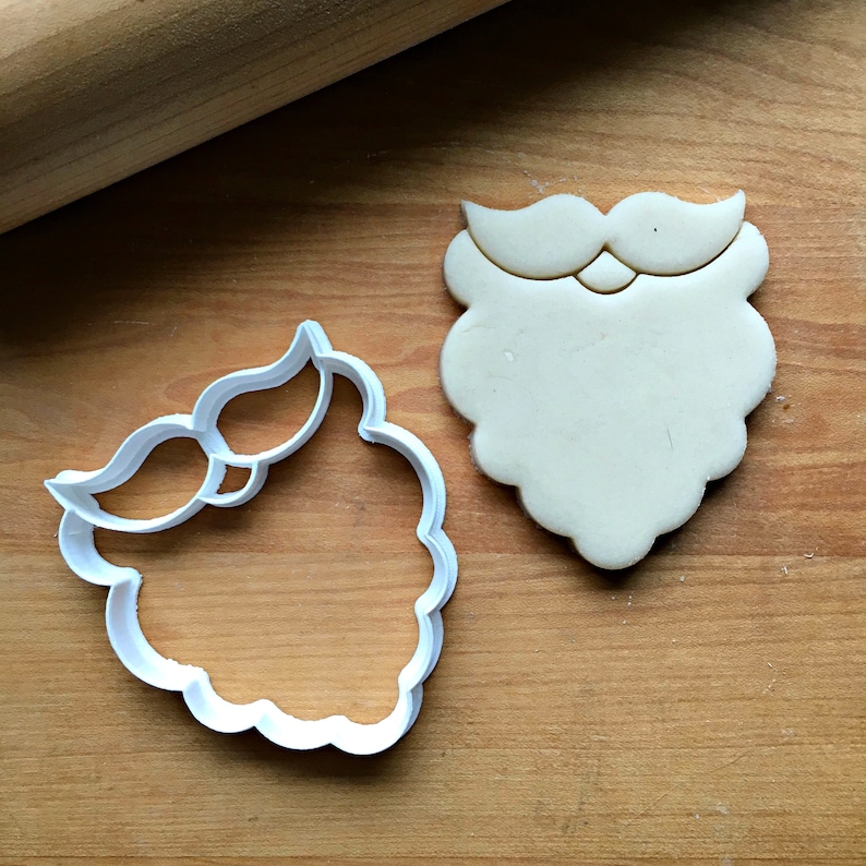 May include: A white plastic Santa beard cookie cutter next to a cookie shaped like a Santa beard. The cookie cutter is on a wooden surface with a rolling pin in the background. The cookie is beige.