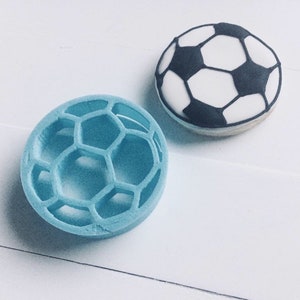 May include: A blue plastic cookie cutter shaped like a soccer ball, next to a decorated sugar cookie with black and white icing.