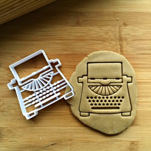 May include: A white plastic cookie cutter shaped like a vintage typewriter, next to a brown cookie dough cutout of the same shape.