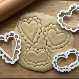 May include: Four white heart-shaped cookie cutters on a wooden surface with a rolling pin and a sheet of cookie dough with three heart-shaped cutouts.
