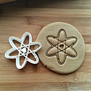May include: A white plastic atom-shaped cookie cutter next to a round of cookie dough with an atom imprint. The cookie cutter has a six-pointed star design with a small circle in the center. The dough is light brown, and the background is a wooden surface.
