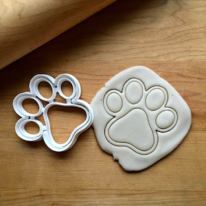 May include: White plastic paw print cookie cutter with a paw print impression in white modeling clay.
