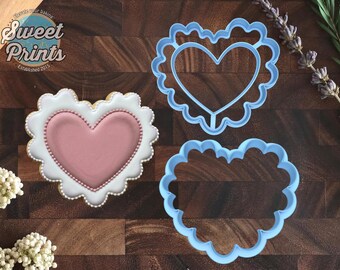 May include: A heart-shaped cookie decorated with white and dusty pink icing, accompanied by two light blue plastic heart-shaped cookie cutters with scalloped edges. The text "Sweet Prints" and "Elevate your baking" is visible in the corner.